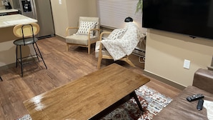 Un salón moderno con una mesa de centro de madera, dos sillones, un sofá, un televisor de pantalla plana y una zona de cocina con refrigerador y dispensador de agua.