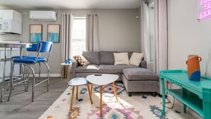 Een moderne woonkamer met een grijze bank, twee kleine ronde tafels, een blauwe stoel, een keuken met een magnetron, en een neonbord aan de muur.