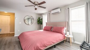 Een slaapkamer met een bed, een plafondventilator, een airconditioningapparaat, een ronde spiegel, een kamerplant en een raam met gordijnen.