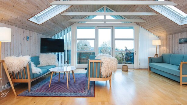 A spacious loft with a high ceiling, wooden walls, and large skylights. There is a cozy seating area with a sofa and chairs, a flat-screen TV mounted on the wall, and a large window offering a view of the outdoors. - 6 Person Holiday Home in Glesborg-by Traum (Glesborg)