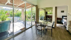 Ein geräumiges Zimmer mit einer großen Glasschiebetür, die auf eine Terrasse führt, einem Essbereich mit Tisch und Stühlen, einem Bett und einer kleinen Küchenzeile.