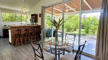 Un vaste espace de salle à manger avec une table et des chaises en verre, une cuisine à l’arrière-plan et de grandes fenêtres offrant une vue sur la verdure.