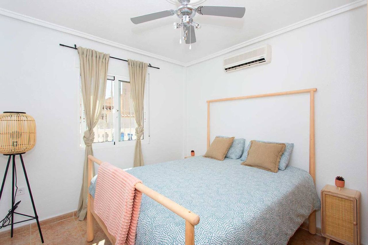 Ein Schlafzimmer mit Bett, Deckenventilator, Klimaanlage, Fenster mit Vorhängen und einem kleinen Nachttisch.