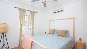 Quarto com cama, ventilador de teto, aparelho de ar condicionado, janela com cortinas e uma mesinha de cabeceira.