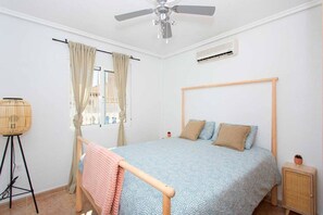 A bedroom with a bed, a ceiling fan, an air conditioning unit, a window with curtains, and a small bedside table.