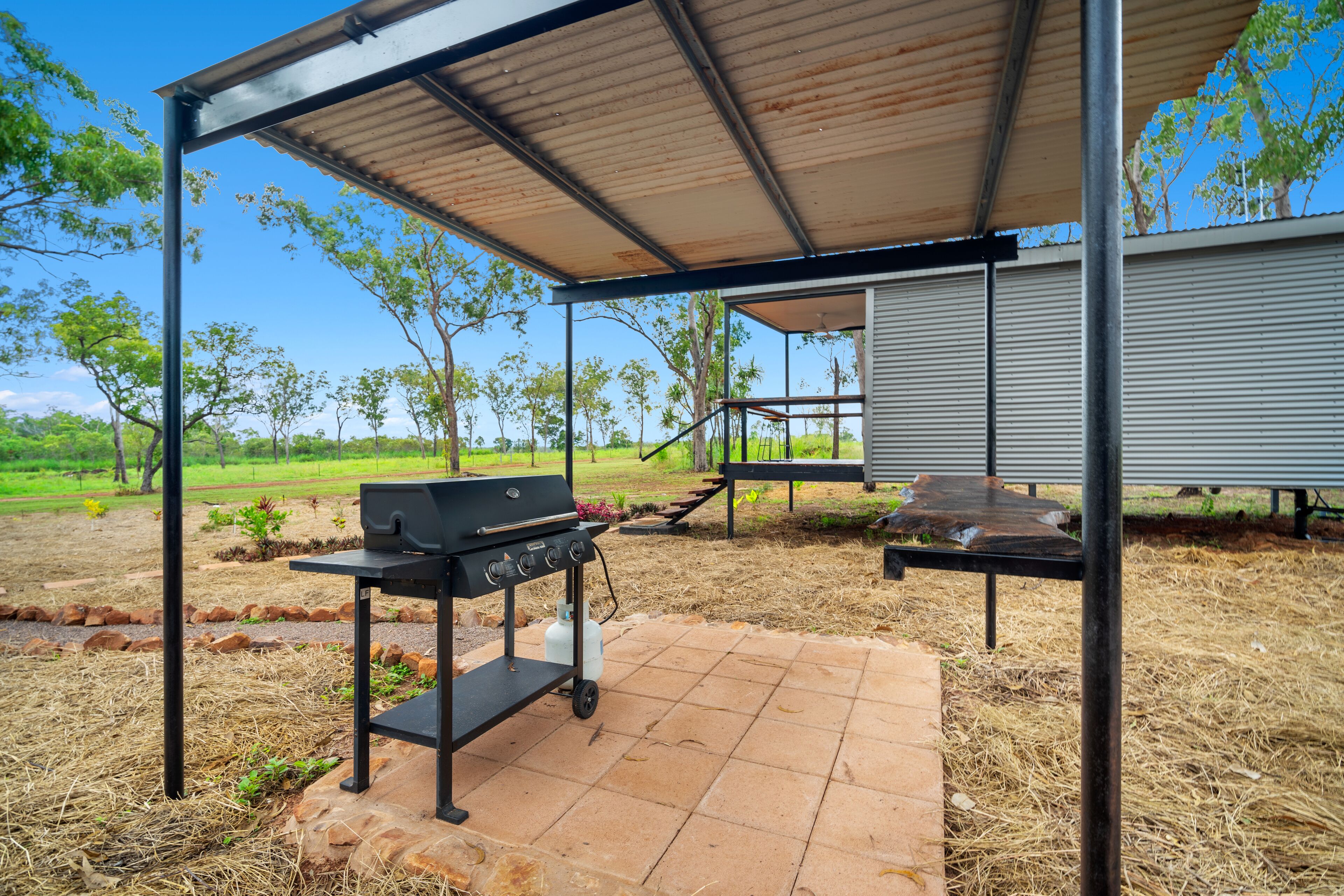 An outdoor patio area with a grill and a barbecue table, surrounded by a metal pergola and a view of a grassy field.