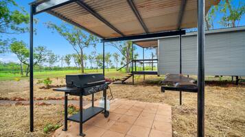 An outdoor patio area with a grill and a barbecue table, surrounded by a metal pergola and a view of a grassy field.