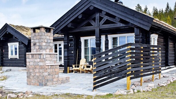 Une cabane en rondins avec une cheminée en pierre, une terrasse en bois et une chaise.