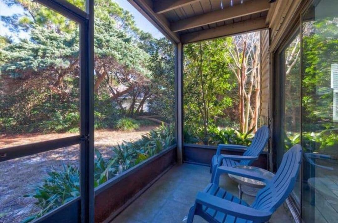 A covered outdoor seating area with chairs and a table, surrounded by glass walls and a view of a garden with trees and plants.