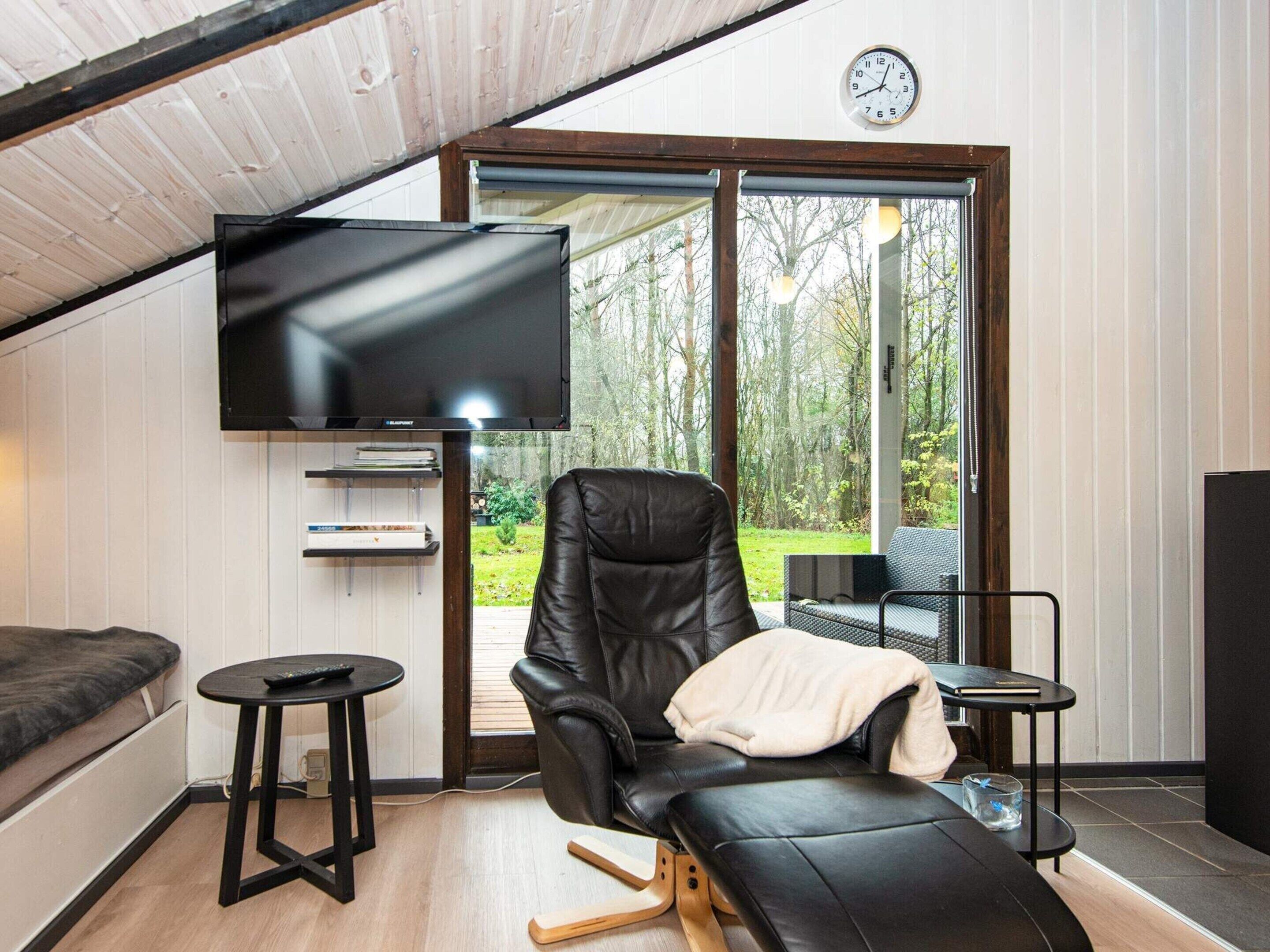 Un salon moderne avec un fauteuil inclinable en cuir, un téléviseur à écran plat et une vue sur un jardin, visible à travers une porte vitrée.