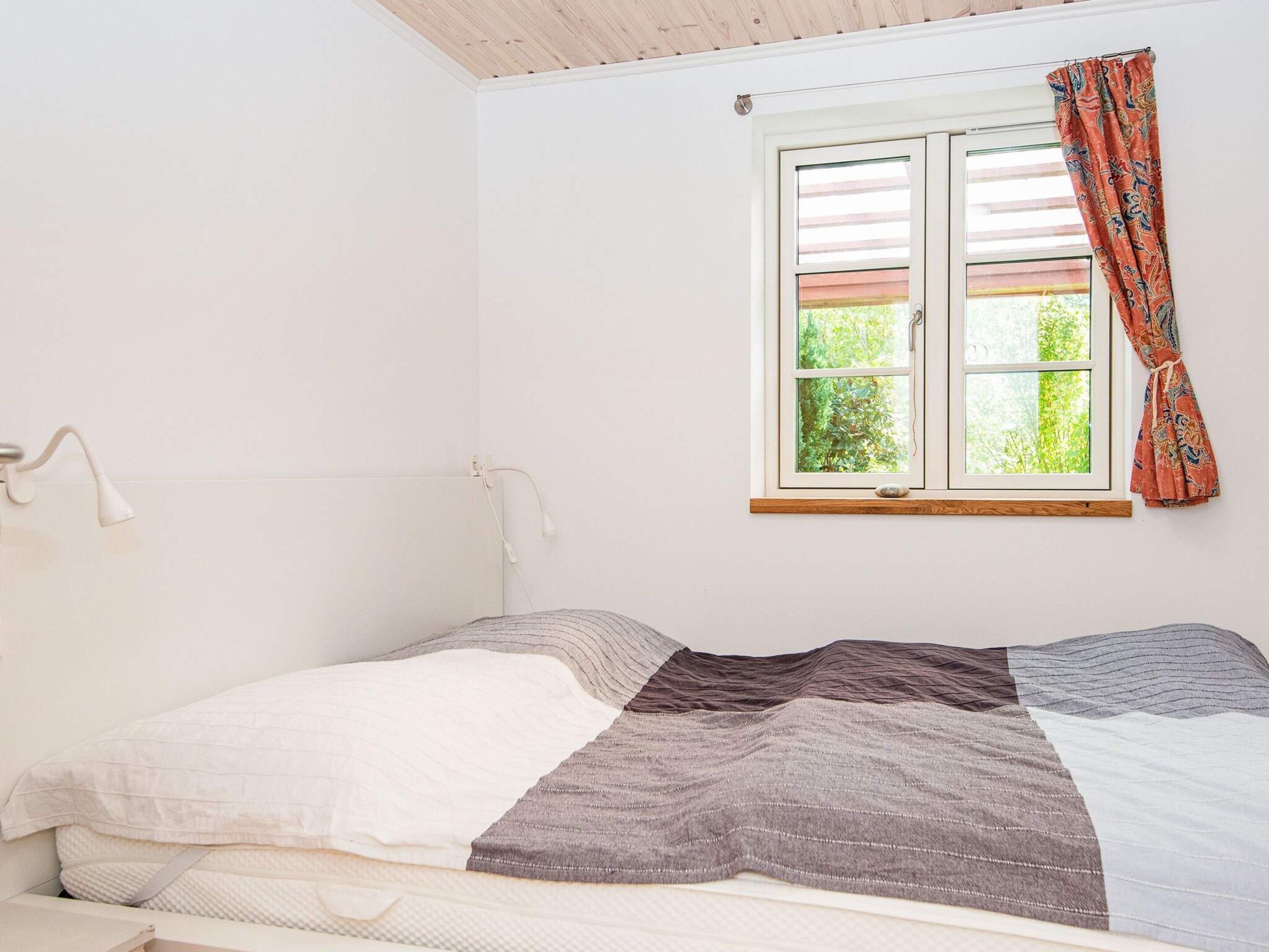 A single-bed room with a wooden ceiling, a window with a view of greenery, and a white bedside lamp.