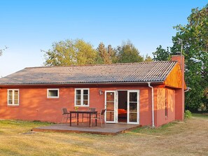 A red brick house with a tiled roof, a wooden deck, and a sliding glass door leading to an interior with a bed and a table. - 6 Person Holiday Home in Ulfborg (Ulfborg)