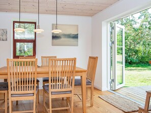 Ein Essbereich mit einem Holztisch und Stühlen, Pendelleuchten, einem Bild an der Wand und Blick ins Grüne durch eine Glastür.