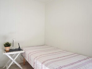 A small, single-bed room with a white wall and a striped bedspread. There is a small table with a potted plant and a lamp.