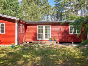 Ett rött trähus med en vit dörr och vita fönster, omgivet av en gräsklädd trädgård och träd.