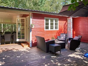 A red wooden building with a covered patio area, furnished with wicker chairs and tables, and a door leading to an outdoor space. - 4 Star Holiday Home in Aakirkeby (Akirkeby)