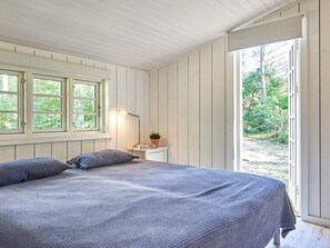 A bedroom with a bed, a nightstand, a lamp, and a window with a view of greenery. - 4 Star Holiday Home in Aakirkeby (Akirkeby)