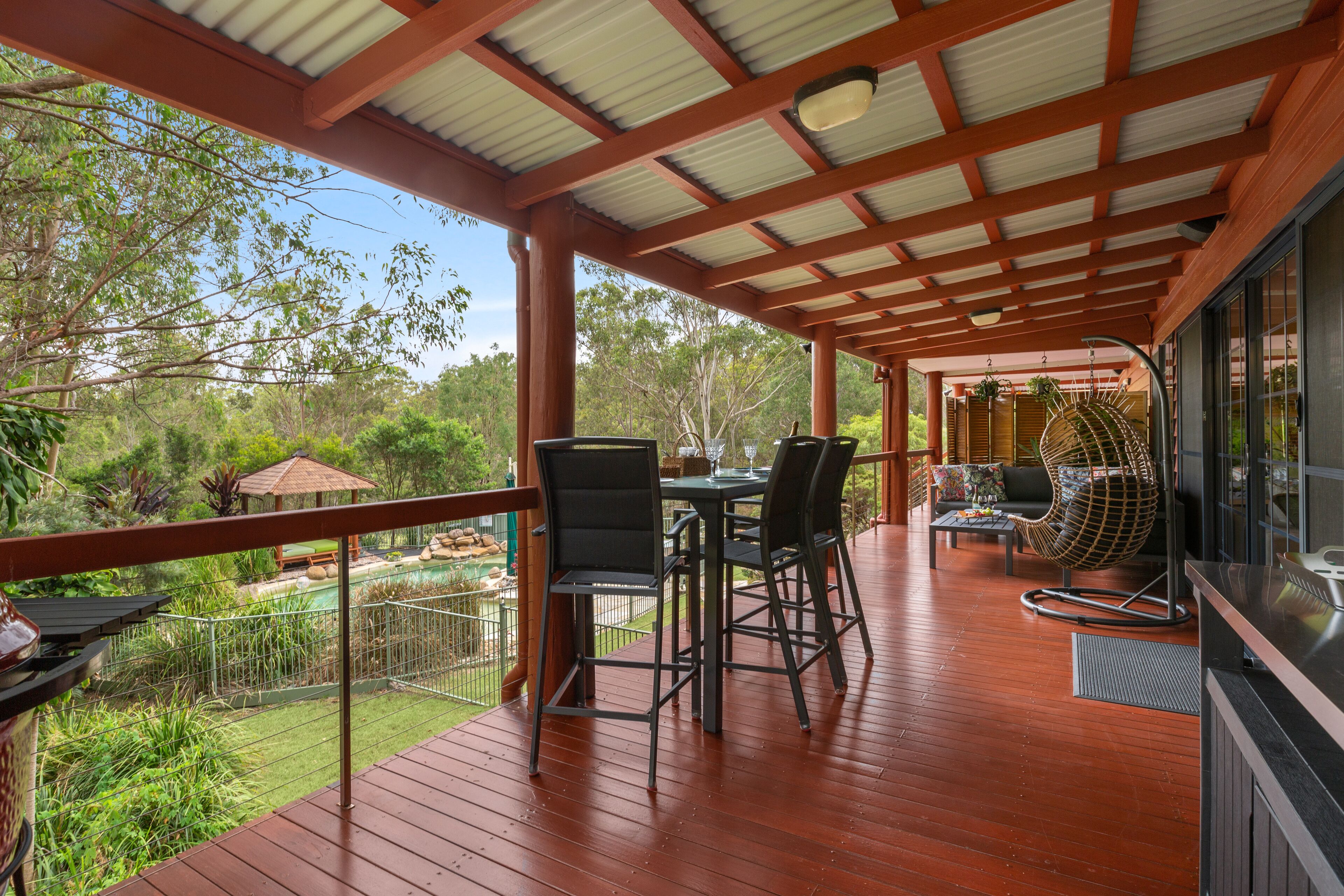 Un espace extérieur abrité doté d’une terrasse en bois, d’une table avec des chaises et d’un coin salon avec un fauteuil rembourré.
