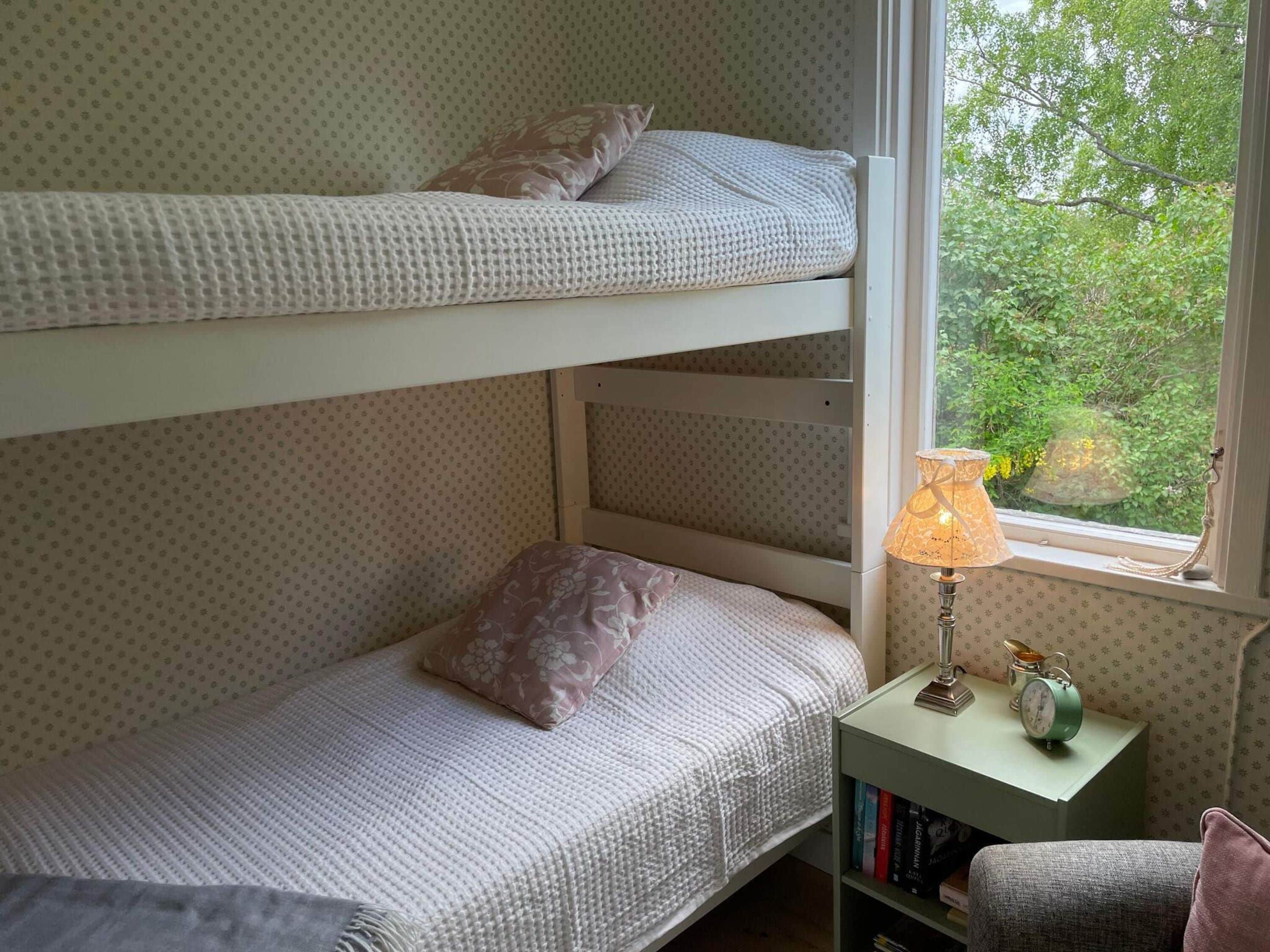A bunk bed room with a window view, a bedside lamp, and a small bookshelf.