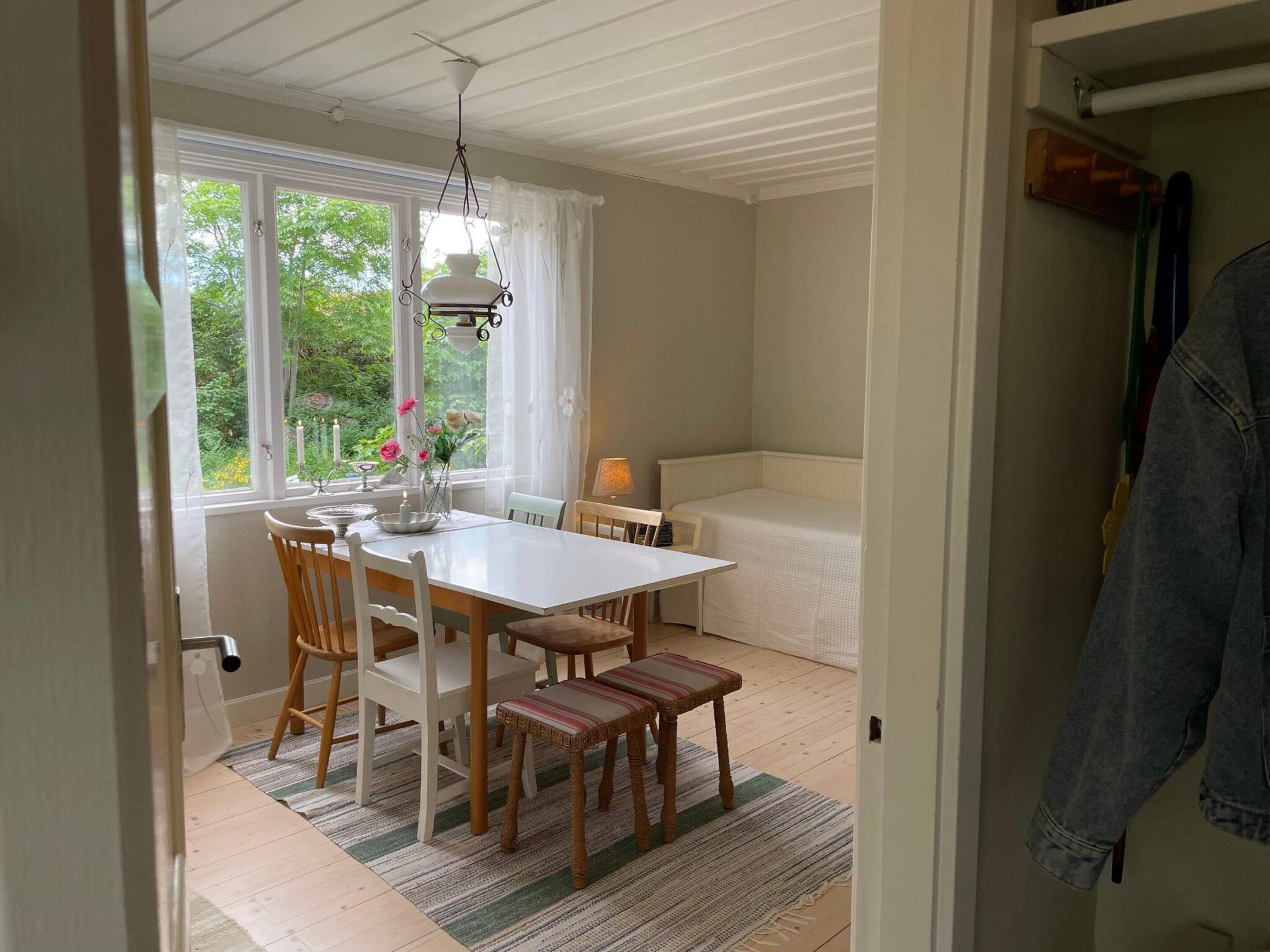 A dining area with a white table, chairs, and a bench. There is a window with curtains, a hanging light fixture, and a view of greenery outside.