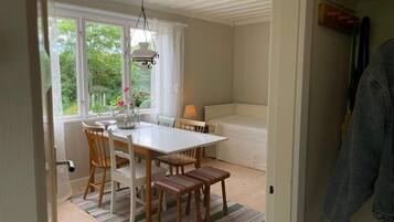 A dining area with a white table, chairs, and a bench. There is a window with curtains, a hanging light fixture, and a view of greenery outside.