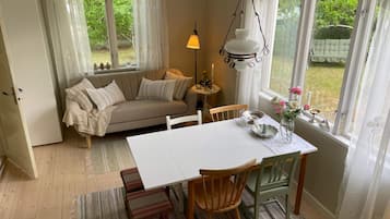 Ett mysigt rum med en soffa, ett matbord och stolar. Det finns ett fönster med gardiner, en hängande lampa och utsikt över omgivningen.