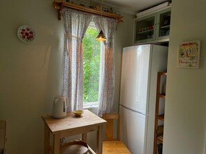 A kitchen with a wooden table, a kettle, a bowl, a refrigerator, and a window with curtains.