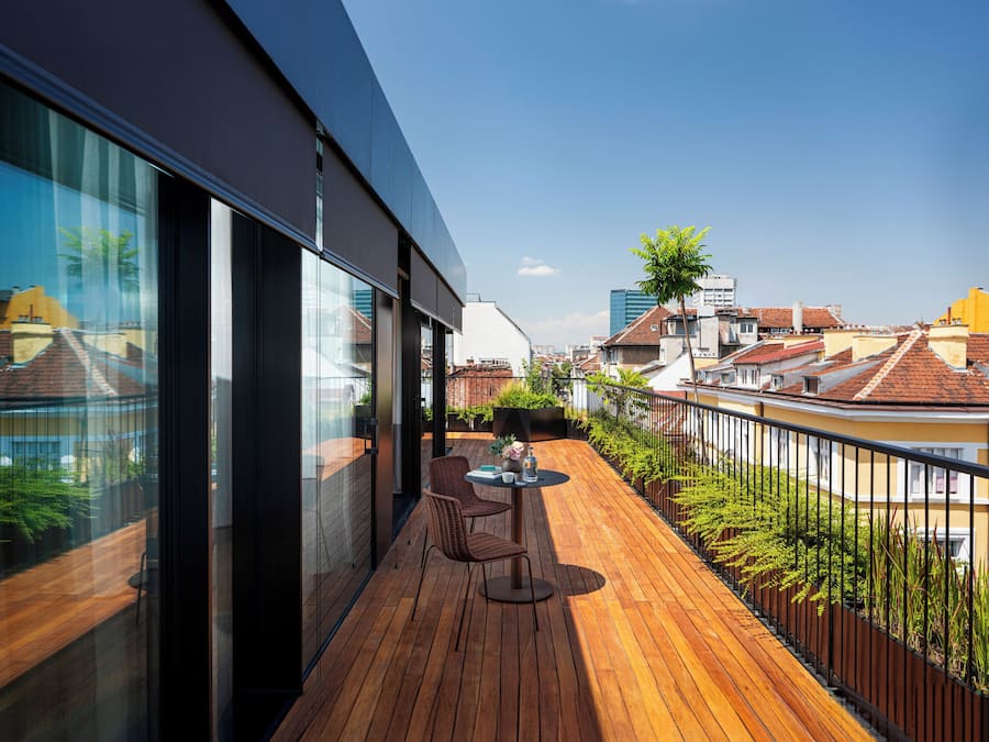 A modern balcony with wooden flooring, a table with chairs, and a glass wall offering a city view.