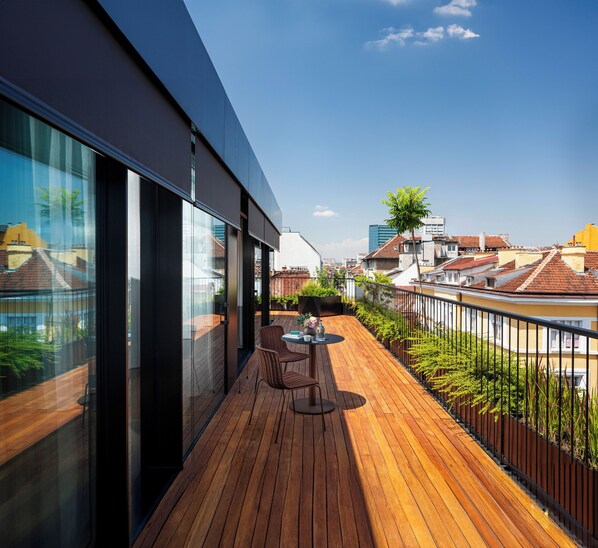 A modern balcony with wooden flooring, a table with chairs, and a glass wall offering a city view.