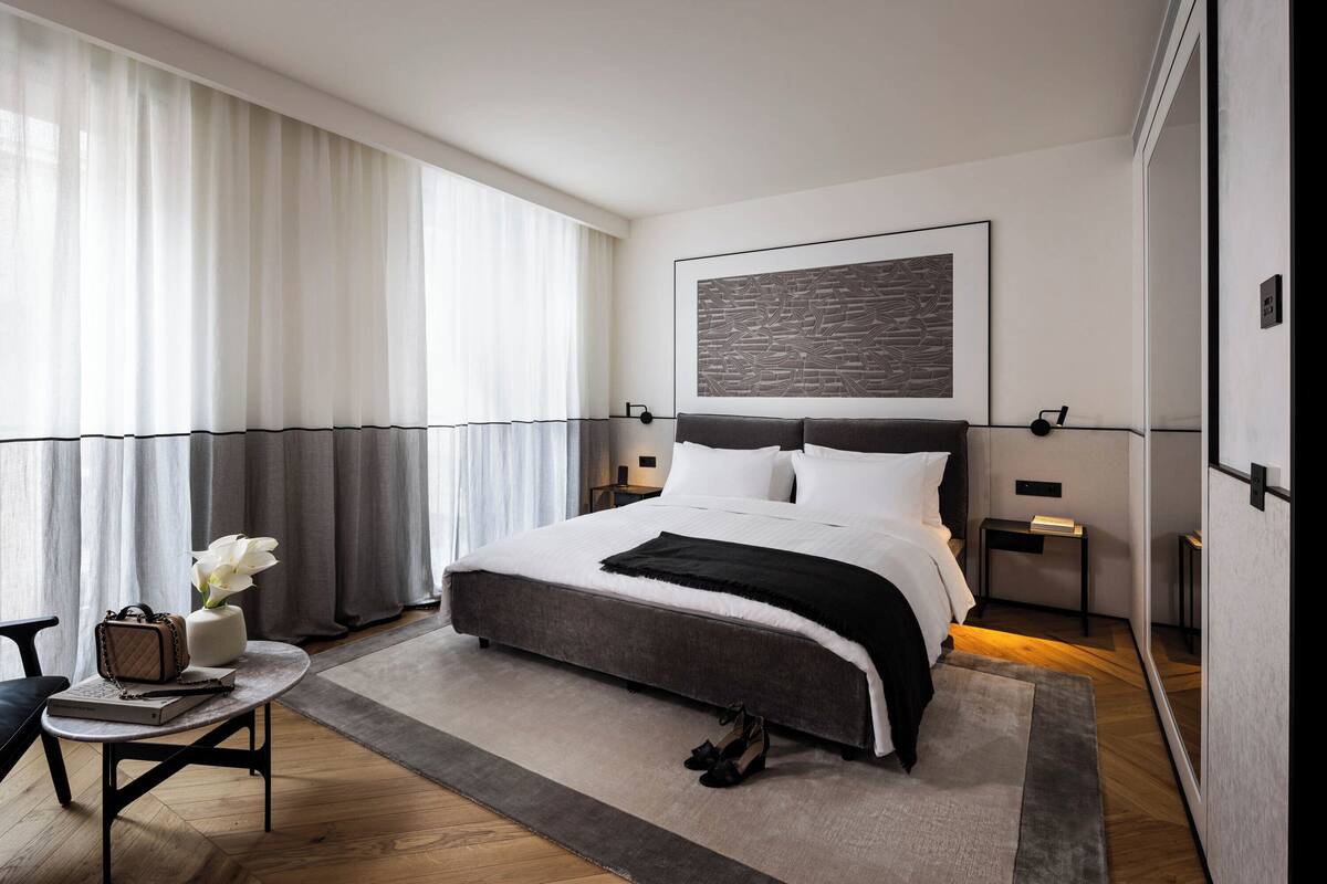 Quarto moderno com uma cama grande, mesa de cabeceira, uma cadeira e uma janela com cortinas.
