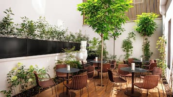 Un patio al aire libre con sillas y mesas de mimbre, rodeado de vegetación y un muro blanco.