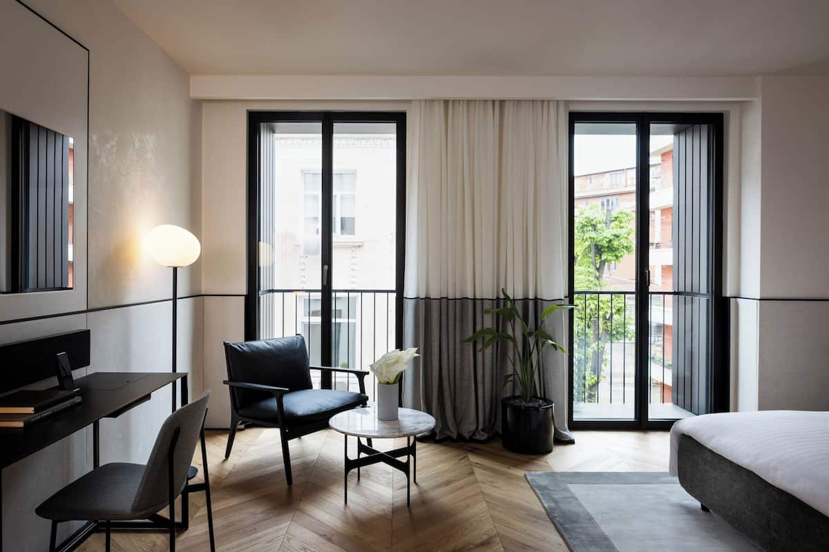 Quarto de hotel moderno com cama, escrivaninha com laptop, cadeira, mesinha com um vaso de flores e janelas amplas com cortinas.