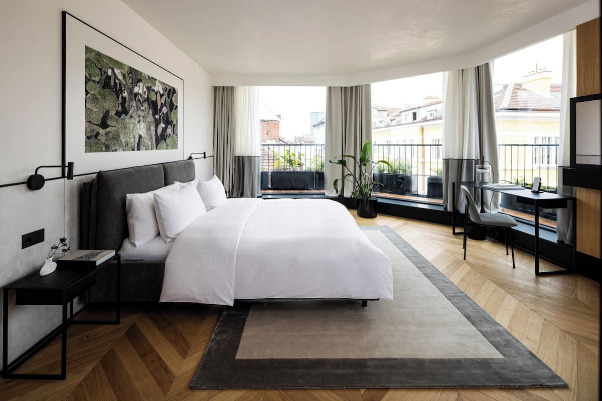 Quarto de hotel moderno com uma cama grande, uma escrivaninha, uma cadeira e uma varanda com plantas.