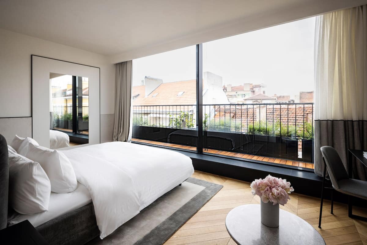 Quarto de hotel moderno com uma cama grande, vista da cidade por uma varanda e uma pequena mesa redonda com um vaso de flores.