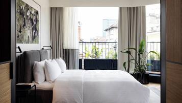 A modern hotel room with a large bed, a nightstand, a potted plant, and a view of the city through large windows.
