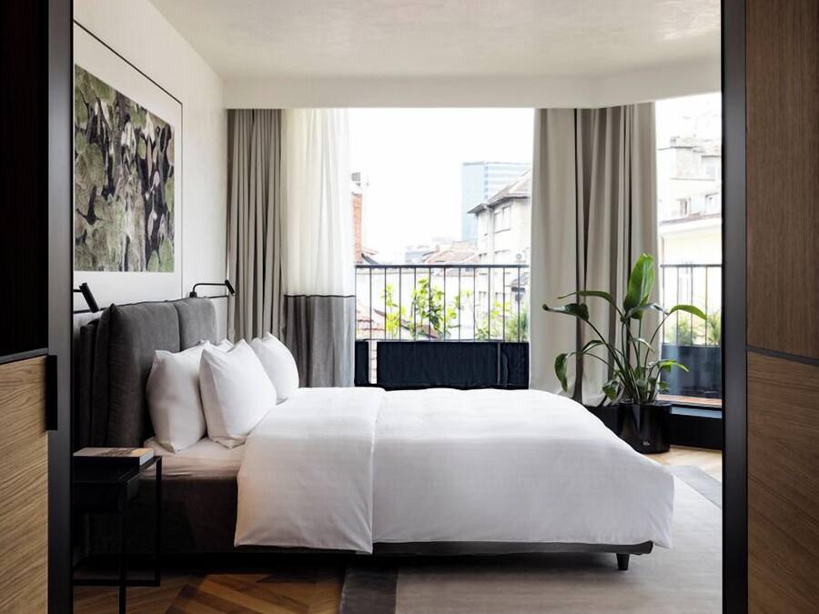A modern hotel room with a large bed, a nightstand, a potted plant, and a view of the city through large windows.