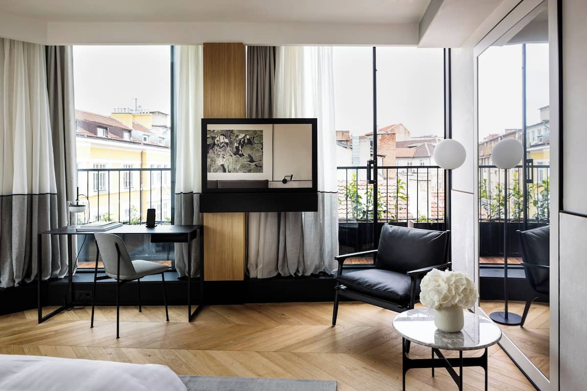 Quarto de hotel moderno com uma janela grande, uma escrivaninha, uma cadeira, um sofá e uma mesinha com um vaso de flores.