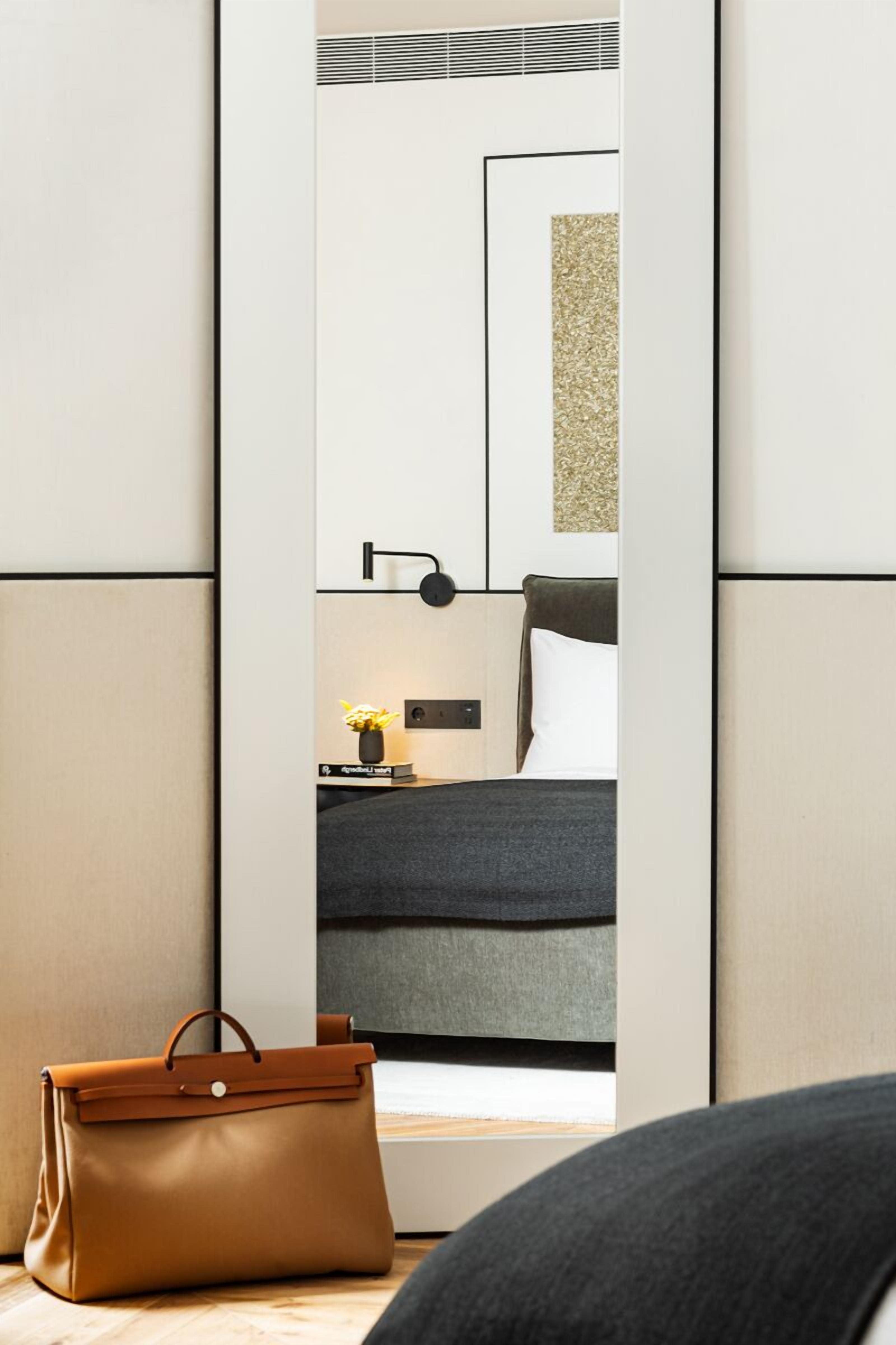 A modern hotel room with a large mirror, a bedside table with a lamp, and a brown leather bag on the floor.