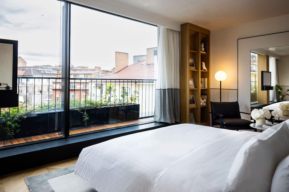 Quarto de hotel moderno com uma cama grande, vista da cidade através de uma varanda, estante e escrivaninha com abajur.