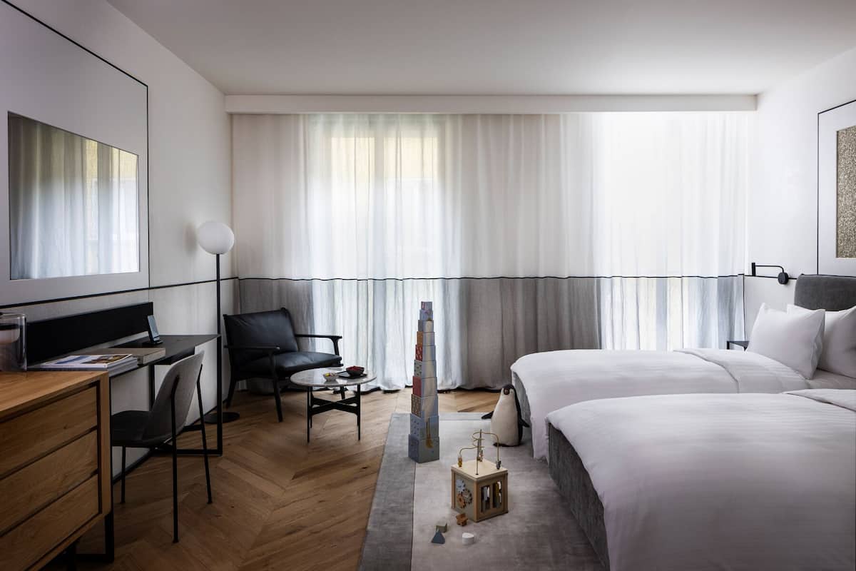 Quarto de hotel moderno com escrivaninha, cadeira, duas camas, mesa pequena com abajur e uma janela grande com cortinas leves.