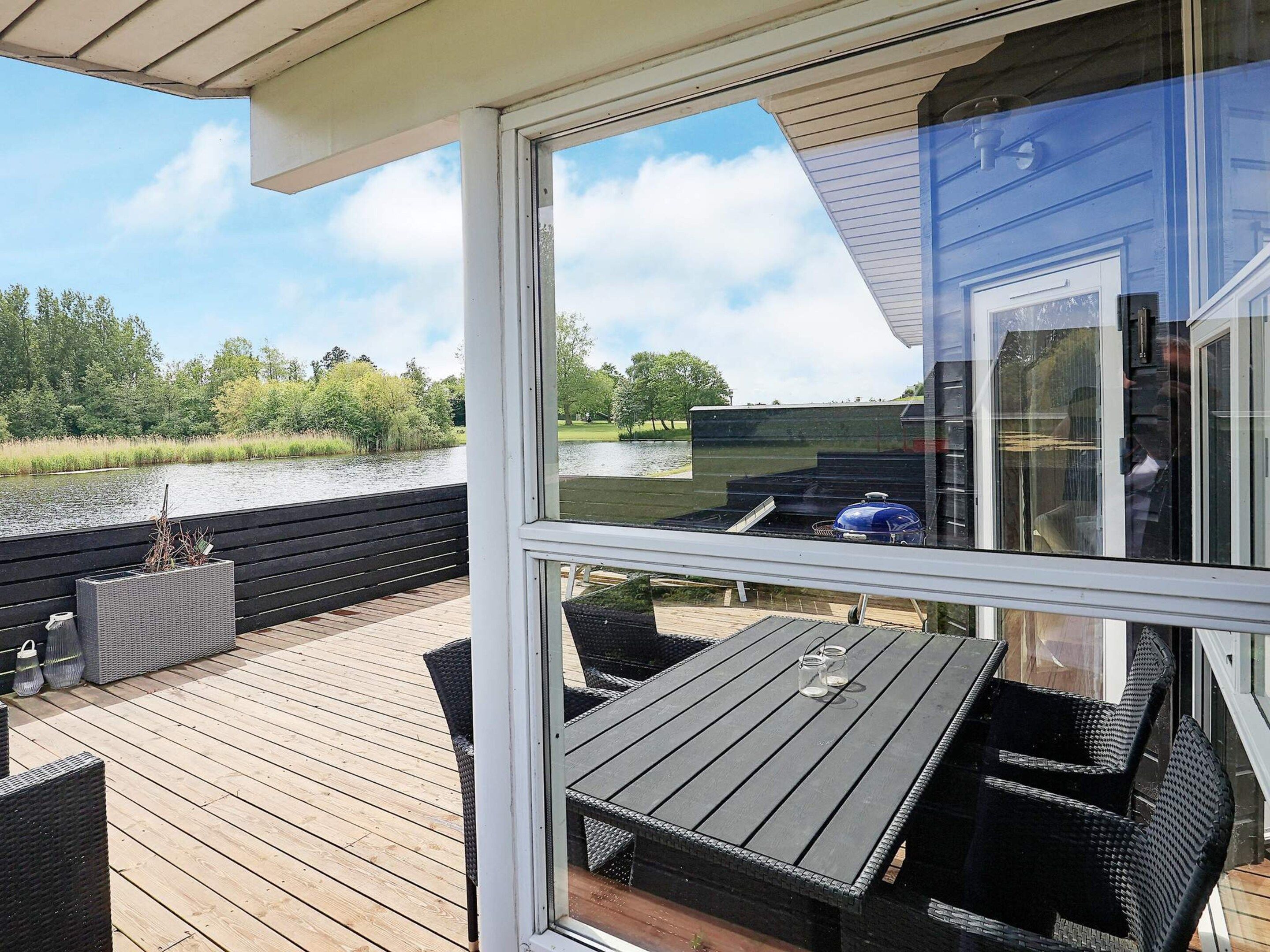 Eine Holzterrasse mit Gartenmöbeln, einer Glastür und Blick auf einen See und viel Grün.