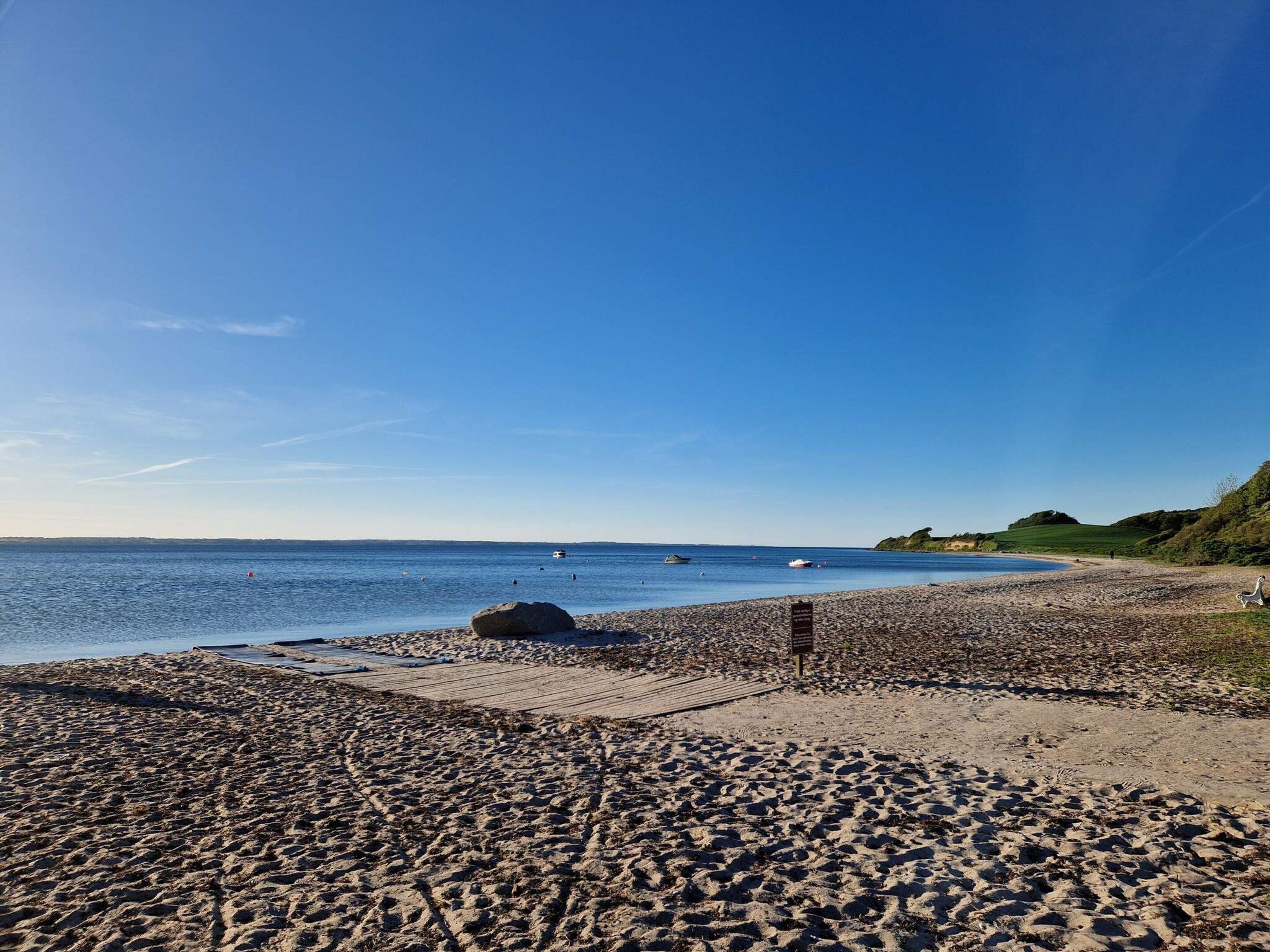 Ein Strand mit einem Kiesweg, der zum Wasser führt, einem Wegweiser und Booten in der Ferne.
