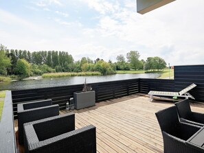 En træterrasse med fletmøbler, udsigt til en sø og en græsplæne med træer.