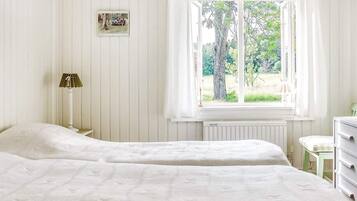 A bedroom with two beds, a window with curtains, a radiator, a lamp, and a small table.
