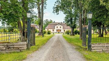 A gravel driveway leading to a building with a traditional facade, flanked by trees and a metal fence with lanterns.