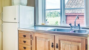 A kitchen with wooden cabinets, a white refrigerator, a stainless steel sink, and a window with a view of a red building and trees. - 3 Person Holiday Home in Flen (Flen)