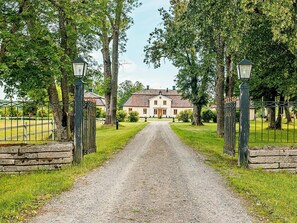 A gravel driveway leading to a large building with a traditional architectural style, surrounded by trees and a metal fence. - 3 Person Holiday Home in Flen (Flen)