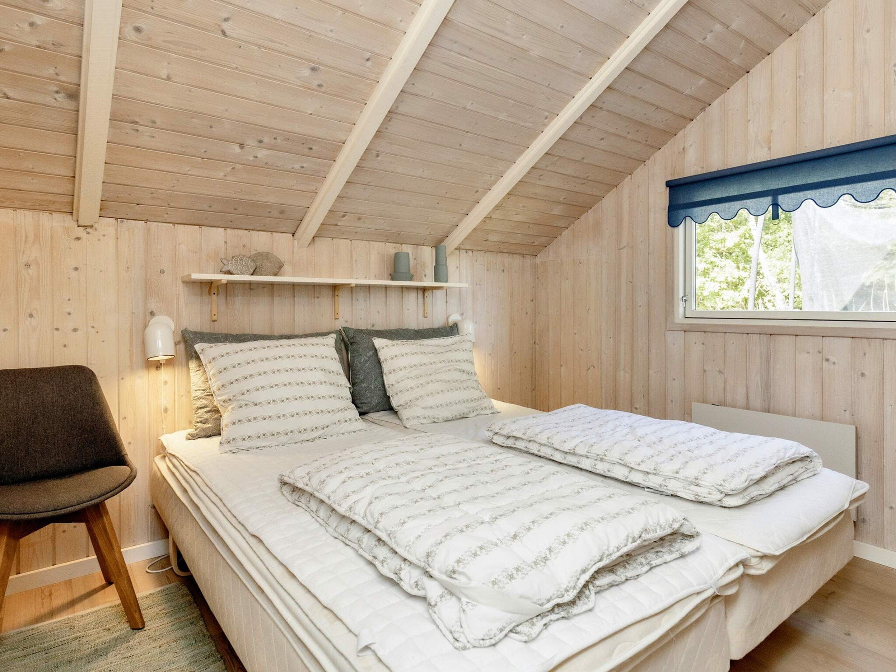 Un dormitorio con techo de madera, cama con ropa de cama blanca, una silla y una ventana con vistas a árboles.