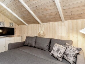Une pièce avec un plafond en bois, un téléviseur à écran plat posé sur un meuble blanc, un canapé gris agrémenté de coussins à motifs floraux et une décoration en macramé accrochée au mur.