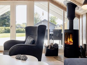 A cozy room with a large window, a comfortable chair, a table, and a wood-burning stove. - 6 Person Holiday Home in Vaeggerlose (Vaeggerlose)